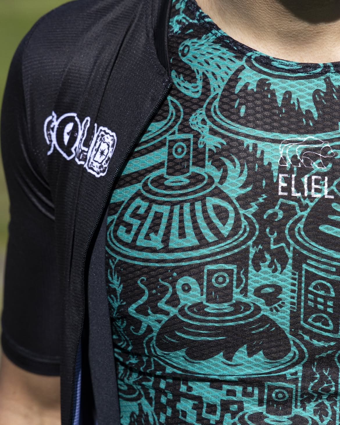 Close-up of a turquoise illustrated Squid cycling jersey beneath a black vest